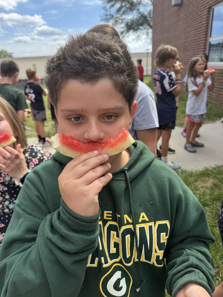 A boy uses a slice of watermelon to hold as a mustaches on his face.