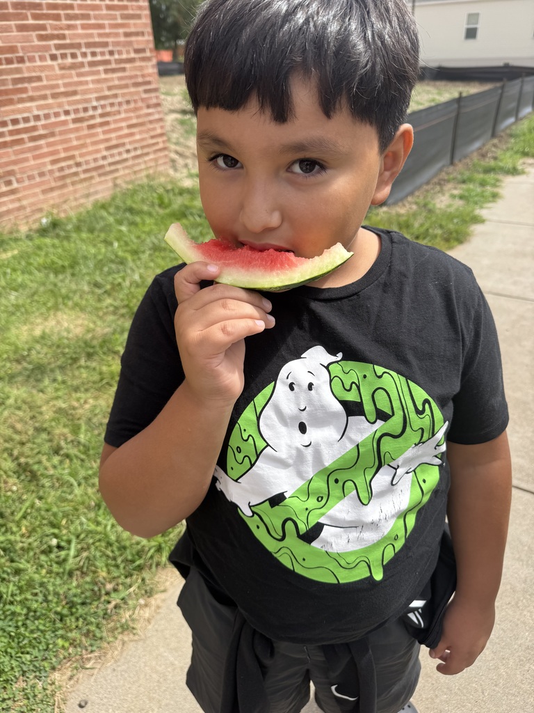 A boy looks at the camera while holding a slice of watermelon up next to his mouth.