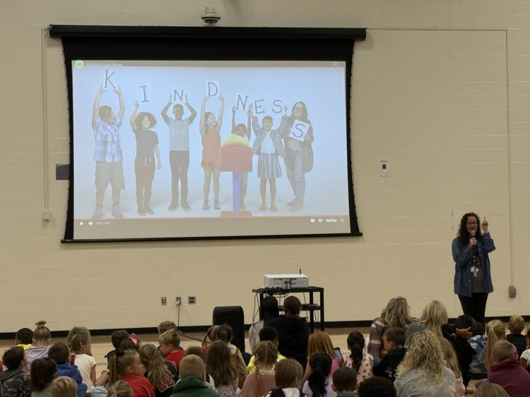 Mrs. Gabriel is standing in front of the gym with a picture of kids holding up letters to spell kindness  
