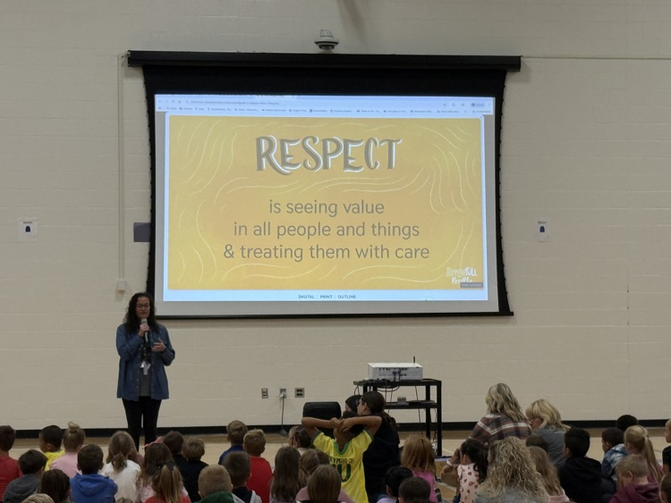 Mrs. Gabriel is standing at the front of the gym with a sign showing Respect - is seeing value in all people and things and treating them with care  