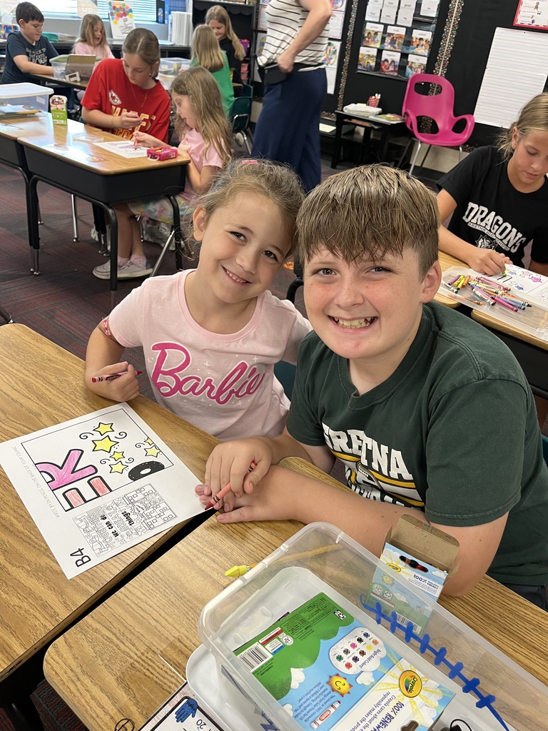 A boy and a girl sitting at a desk with a coloring page in front of them.  They are both smiling.