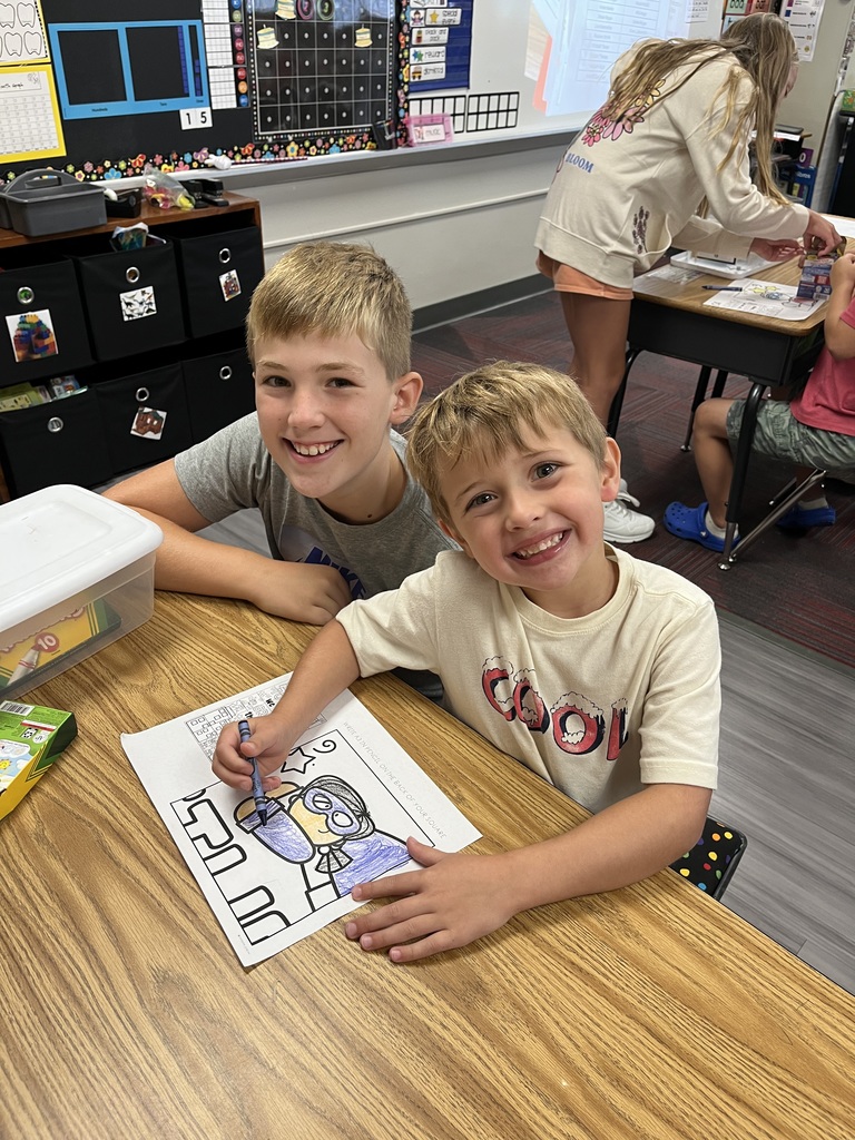 Two boys sitting at a desk with a coloring page in front of them.  They are both smiling.