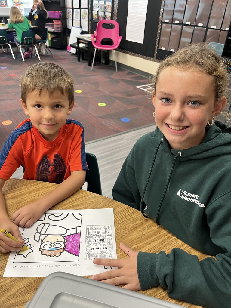 A boy and a girl sitting at a desk with a coloring page in front of them.  They are both smiling.