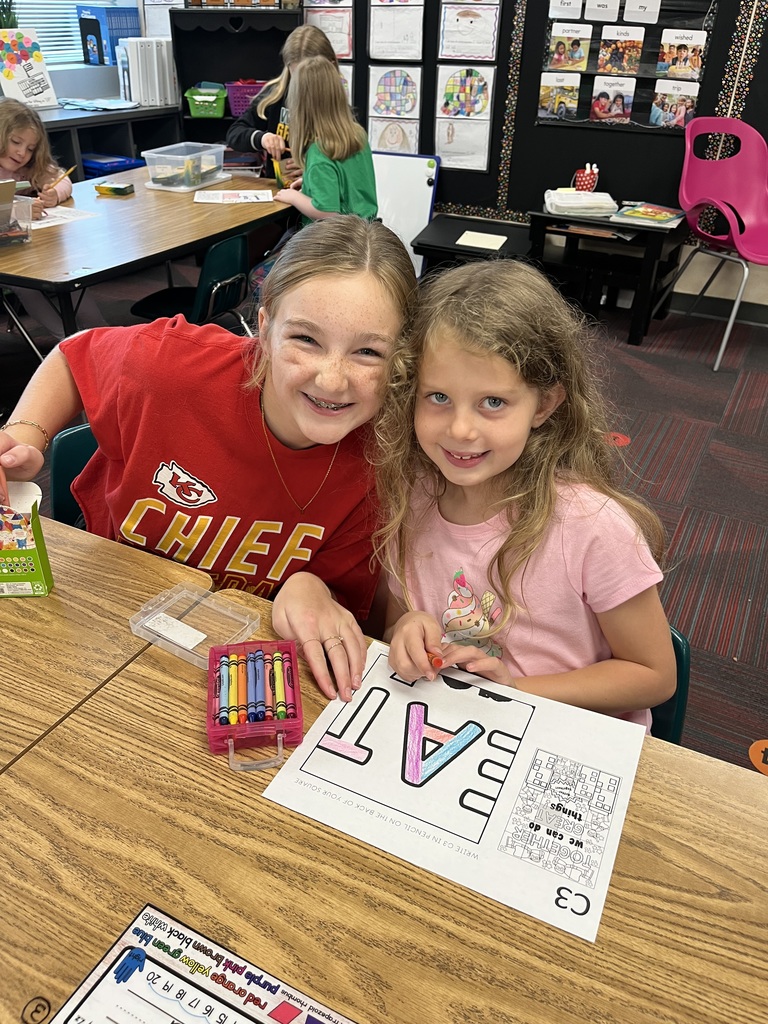 Two girls sitting at a desk with a coloring page in front of them.  They are both smiling.