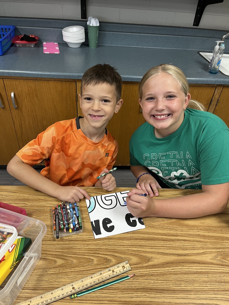 A boy and a girl sitting at a desk with a coloring page in front of them.  They are both smiling.