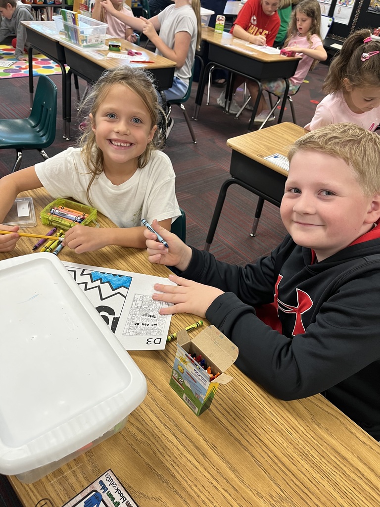 A boy and a girl sitting at a desk with a coloring page in front of them.  They are both smiling.