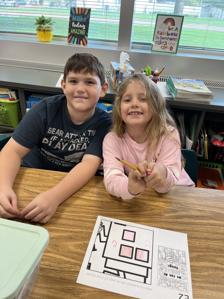 A boy and a girl sitting at a desk with a coloring page in front of them.  They are both smiling.