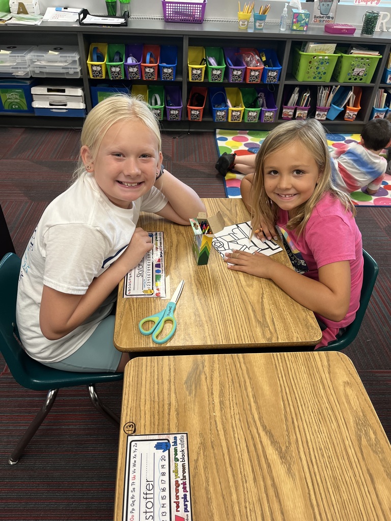 Two girls sitting at a desk with a coloring page in front of them.  They are both smiling.