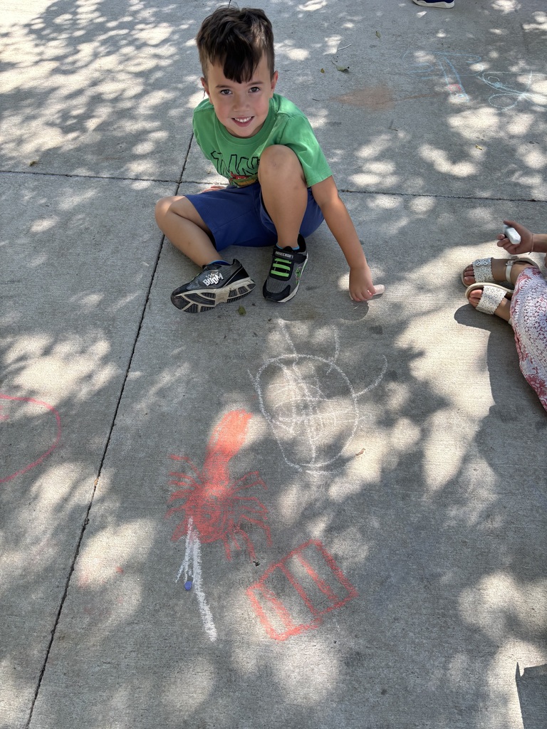 A child sits on the sidewalk while sitting next to the bird he drew with sidewalk chalk.