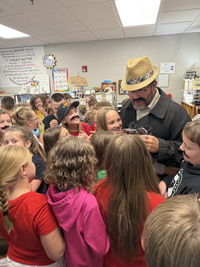 Group of students standing around a man wearing a hat and a fake mustache.