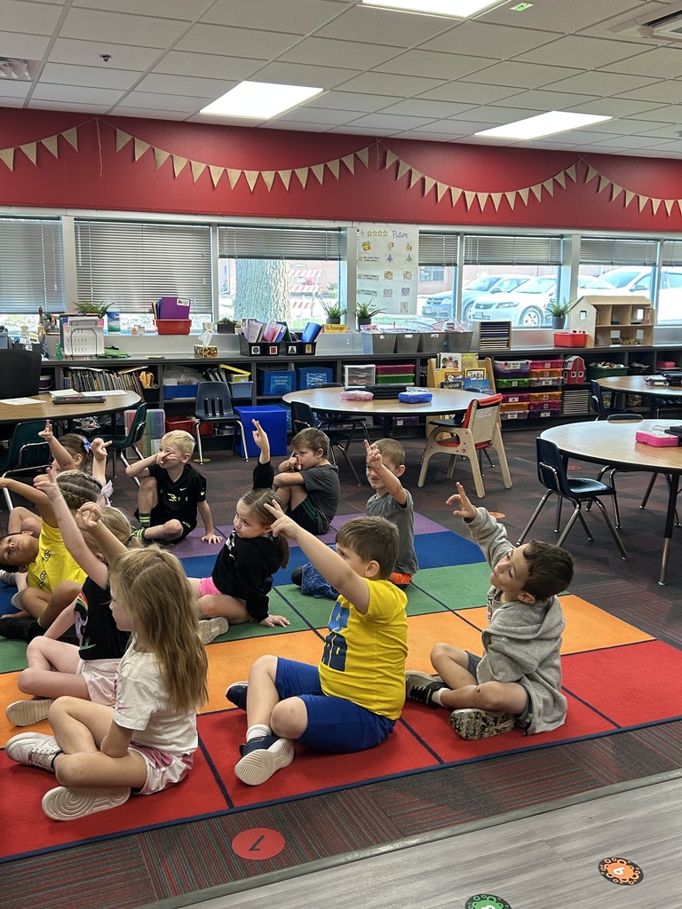 Students sitting on a colored rug with two fingers raised listening intently to someone at front of room.