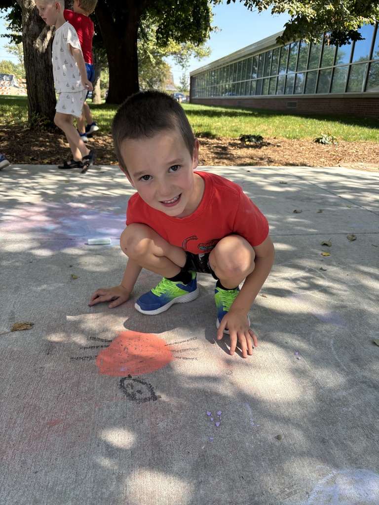 A boy sitting on a sidewalk looking at the camera in front of the sidewalk chalk drawing that he did.