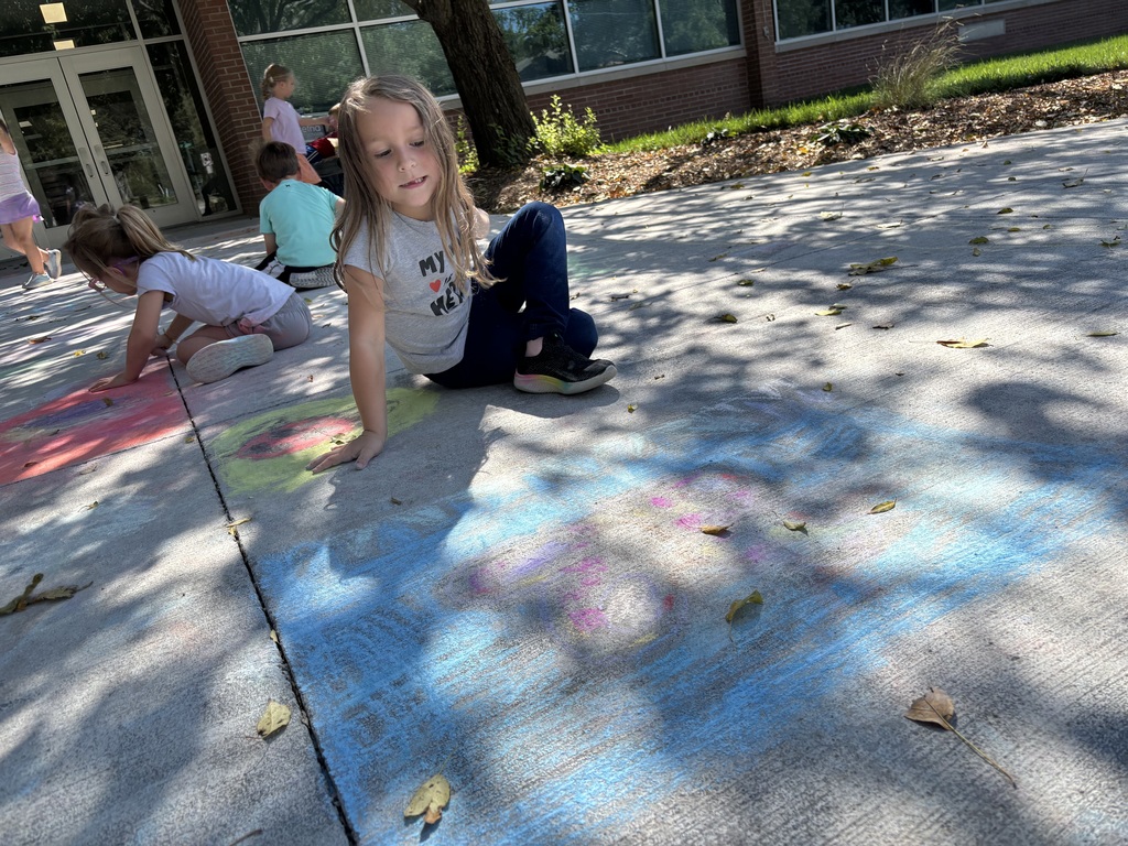 A girl sitting on a sidewalk looking down at the sidewalk chalk drawing that she did.