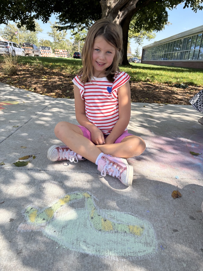 A girl sitting on a sidewalk looking at camera in front of the sidewalk chalk drawing that she did.