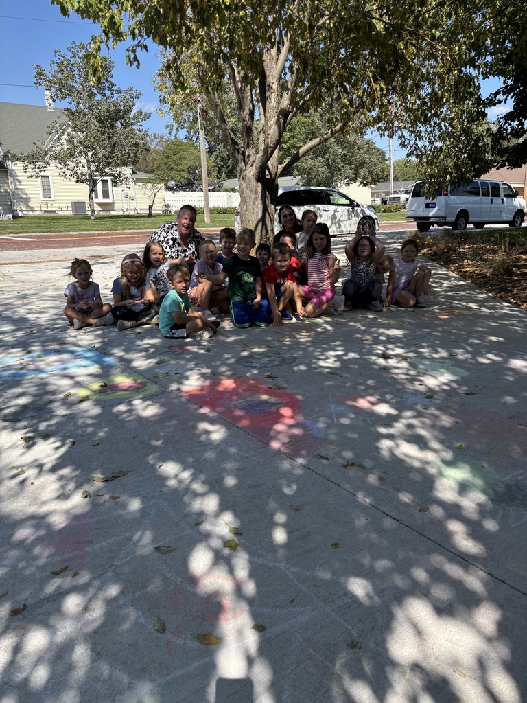 Group of students and two adults sitting on a sidewalk with sidewalk chalk visible on the sidewalk.