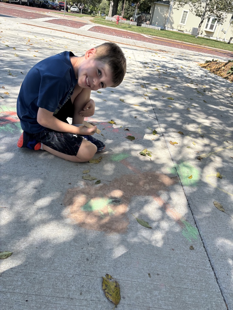 A boy sitting on a sidewalk looking at the camera in front of the sidewalk chalk drawing that he did.