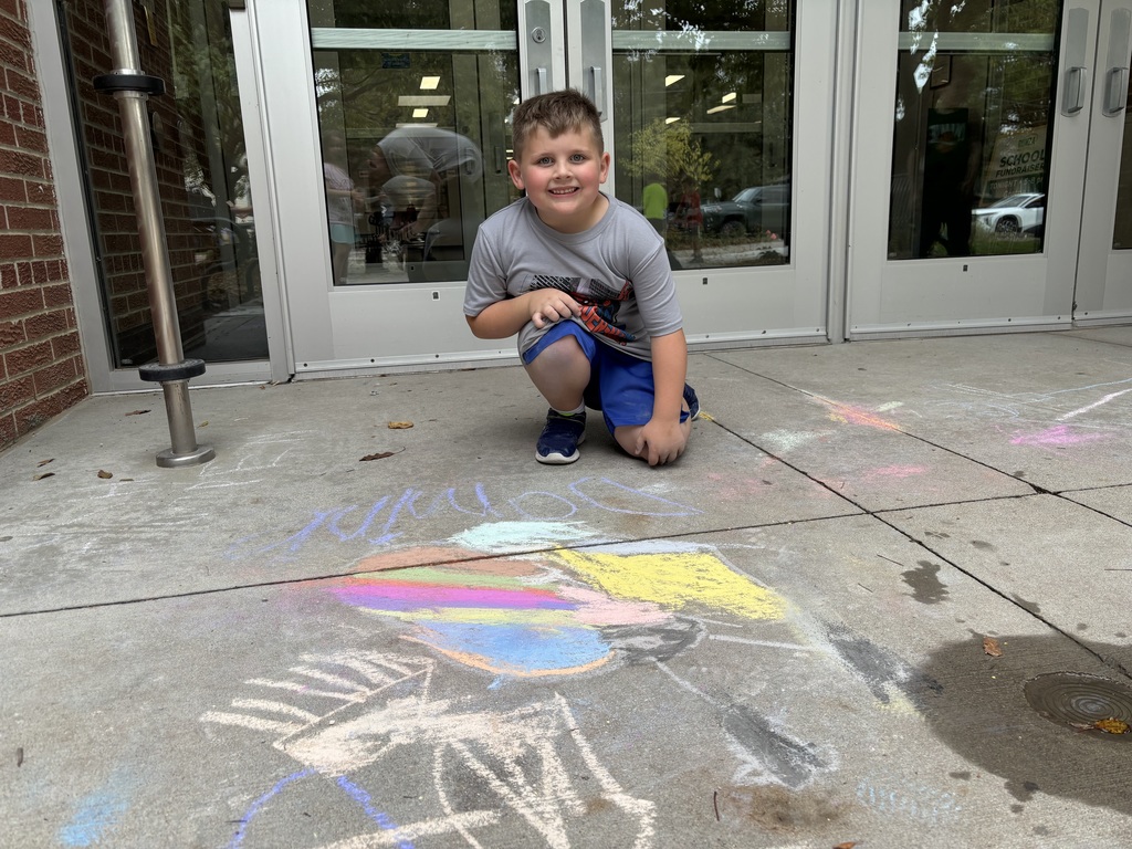 Boy sitting on sidewalk looking at camera in front of drawing made by chalk.