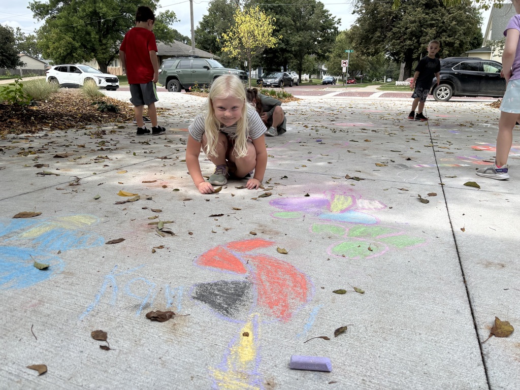 Girl sitting on sidewalk looking at camera in front of drawing made by chalk.
