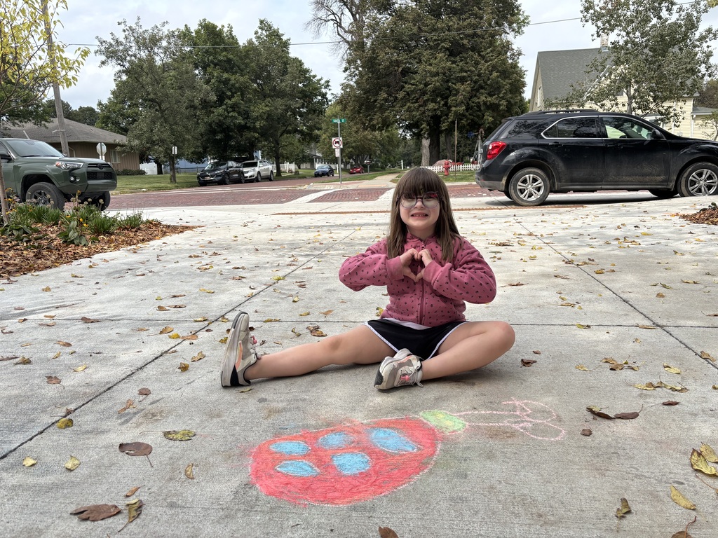 Girl sitting on sidewalk looking at camera in front of drawing made by chalk.