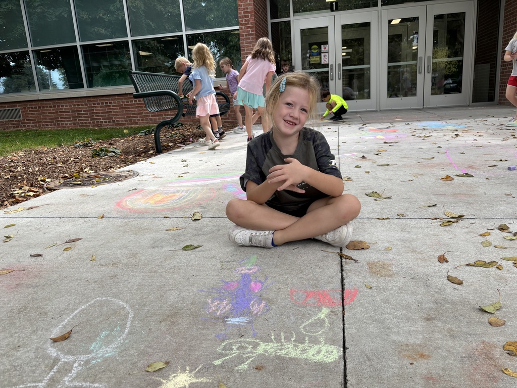Girl sitting on sidewalk looking at camera in front of drawing made by chalk.