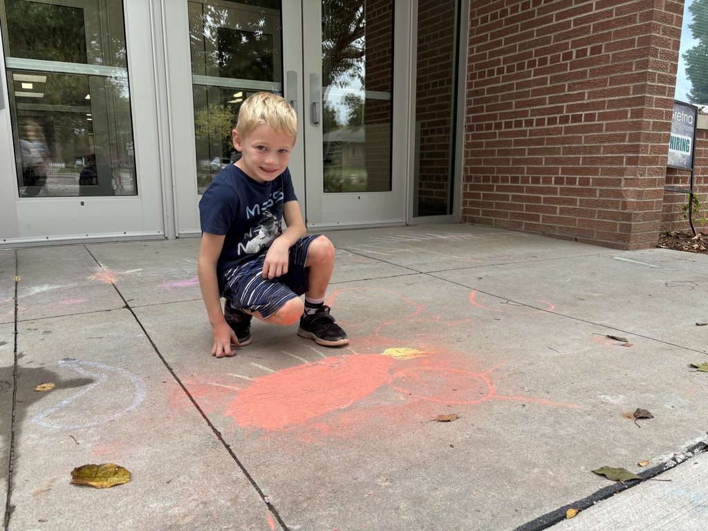 Boy sitting on sidewalk looking at camera in front of drawing made by chalk.
