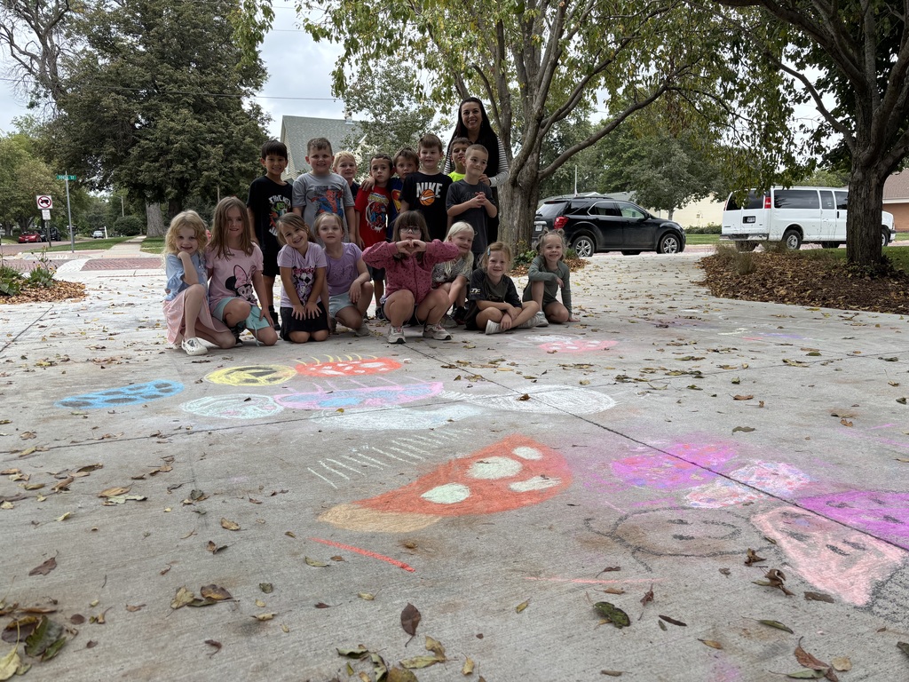 Group of kids, some standing and some sitting with an adult posing for a photo on a sidewalk full of chalk drawings.