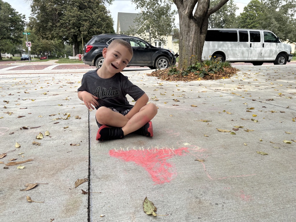 Boy sitting on sidewalk looking at camera in front of drawing made by chalk.