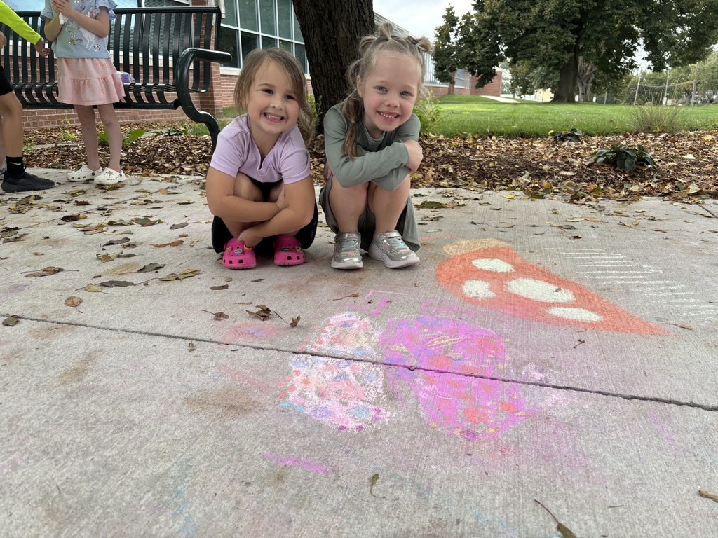 Two girls sitting on sidewalk looking at camera in front of drawing made by chalk.