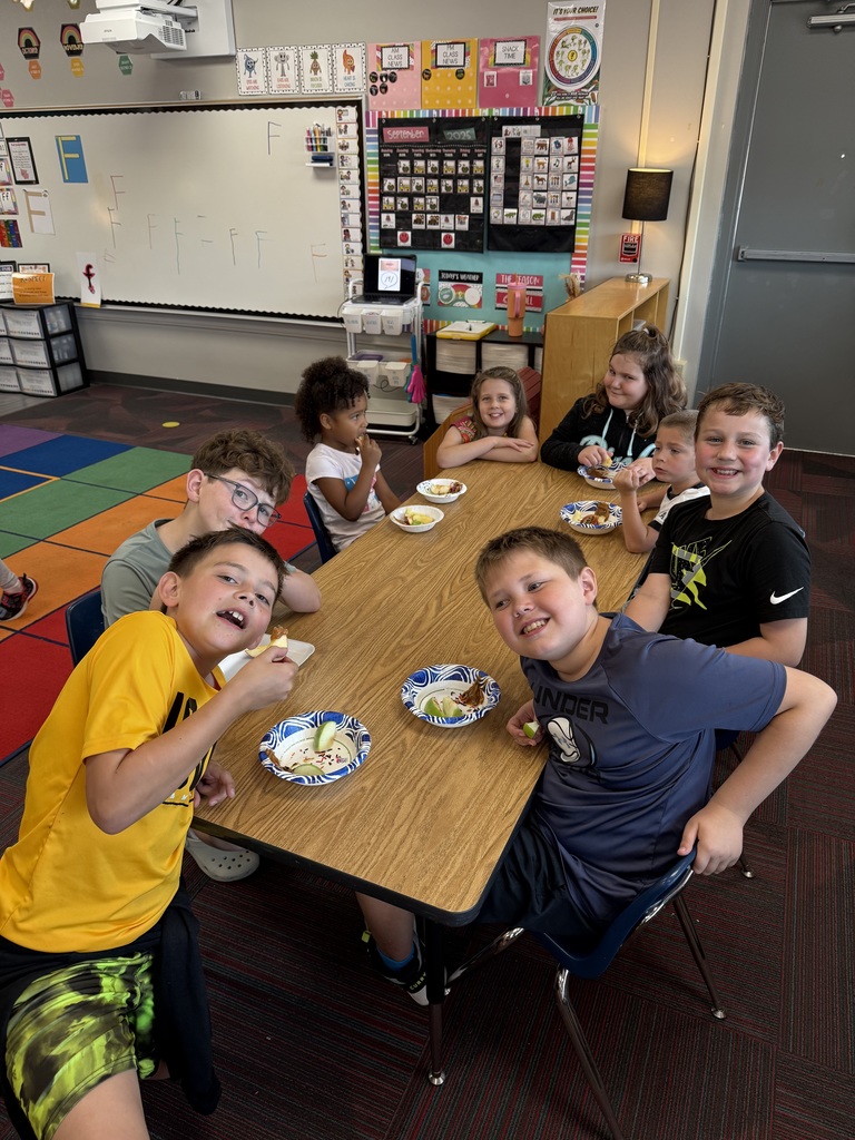 Group of students sitting around a large table with books and apples in front of them smiling.