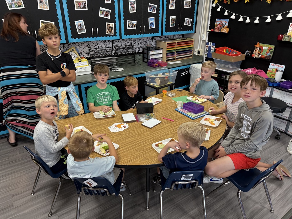 Group of students sitting around a large table with books and apples in front of them smiling.