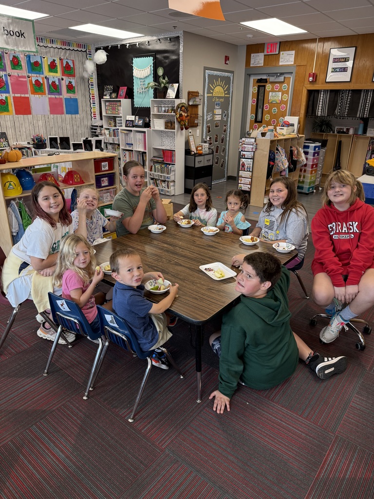 Group of students sitting around a large table with books and apples in front of them smiling.