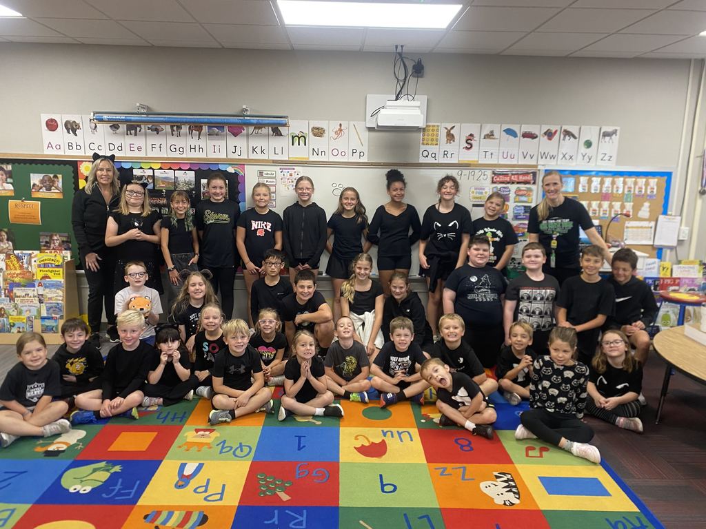 Group of students and their teachers wearing black.  Some are sitting on a colored carpet while others are standing.  