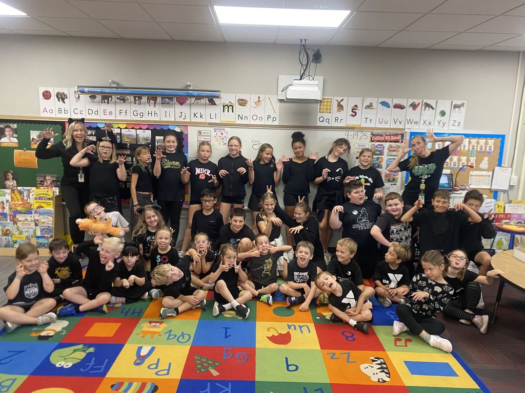 Group of students and their teachers wearing black.  Some are sitting on a colored carpet while others are standing.  Students are making silly faces.