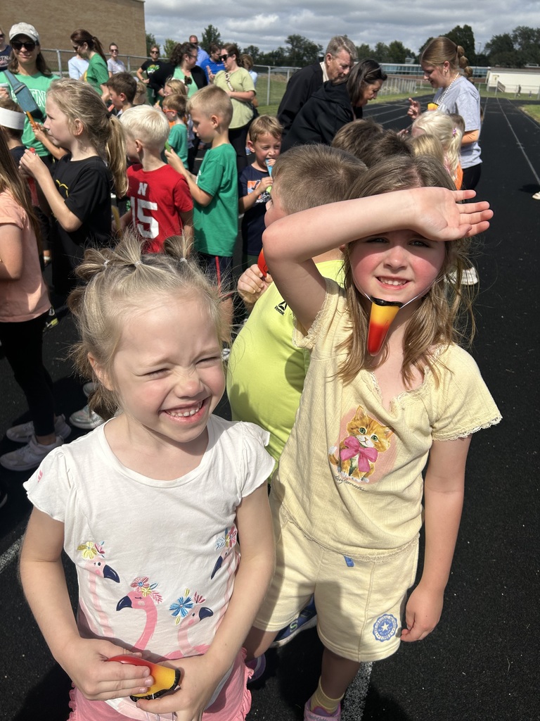 Two girls smiling, one wearing a toucan beak around her chin looking at the camera shielding their eyes from the sun.