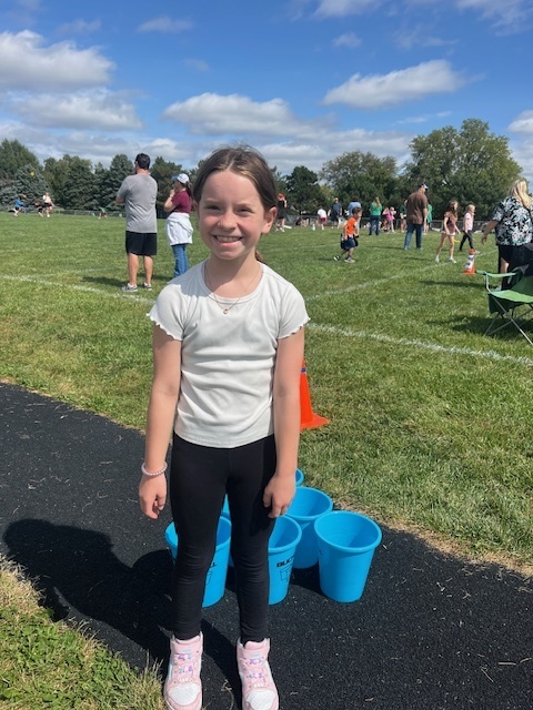A girl smiling while standing in front of blue buckets and a grass field.