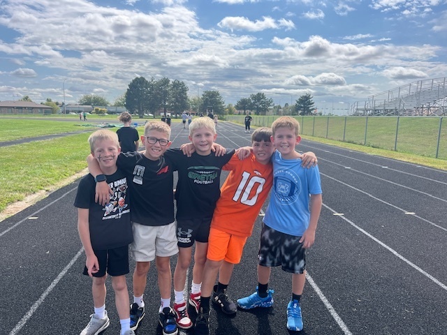 Five boys smiling with their arms wrapped around each other on a running track.