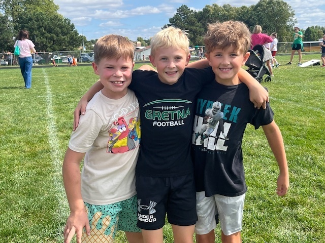 Three boys smiling with their arms wrapped around each other on a grass field.