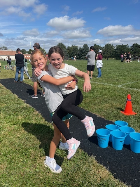 A girl holds up another girl in front of a grass field while smiling.