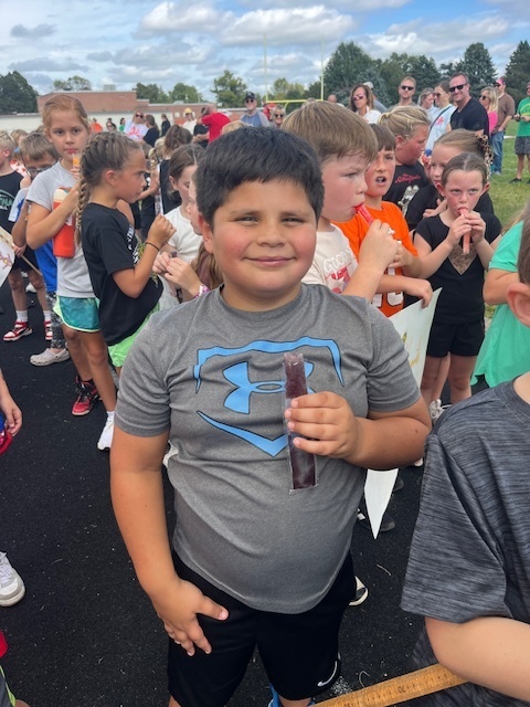 A boy smiles while holding a popsicle with other kids in the background eating popsicles.