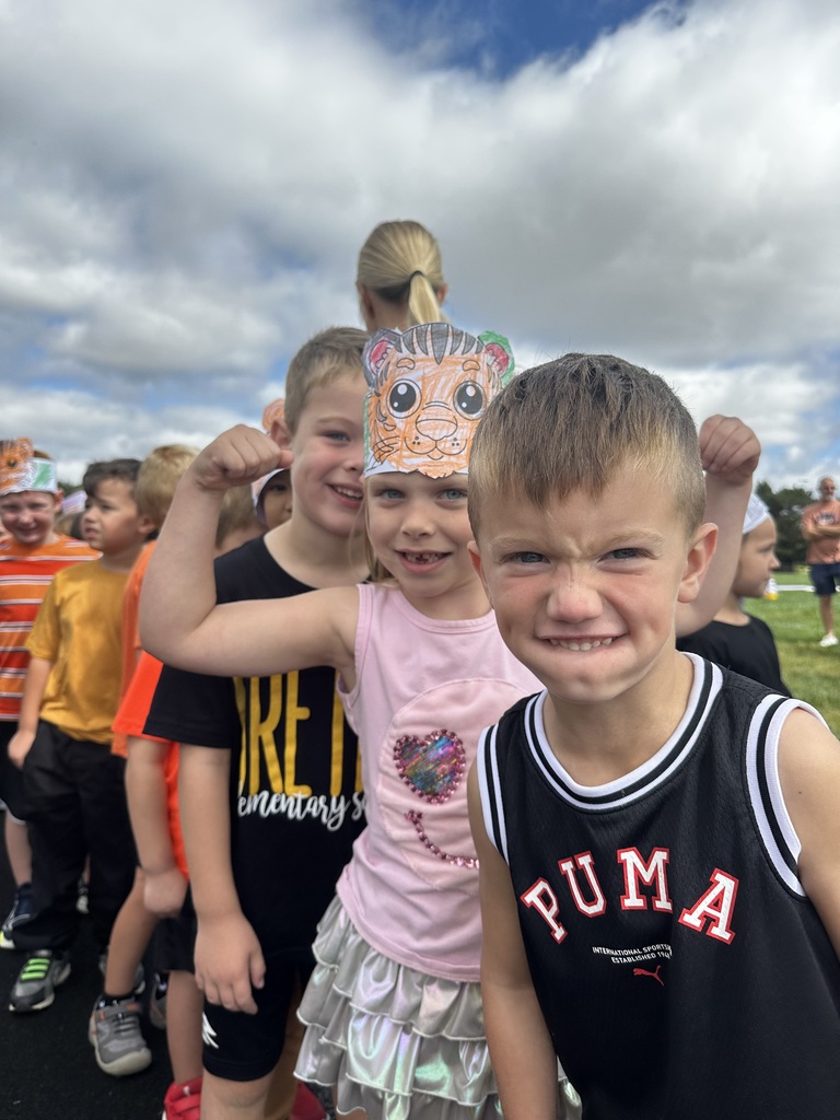 Three students stand for a picture while smiling. One girl holds her arms up to show off her muscles while wearing a tiger headband.