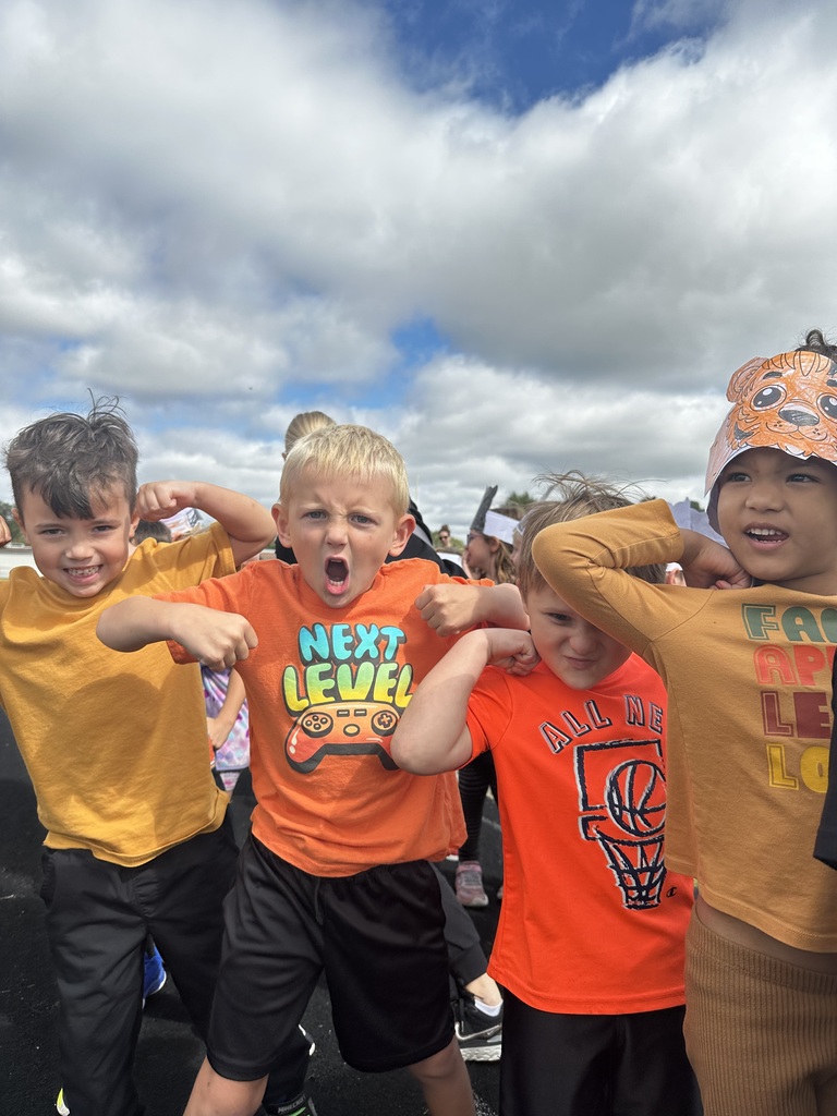 Four students standing while wearing orange and yellow t shirts while holding up their arms to show off their muscles.