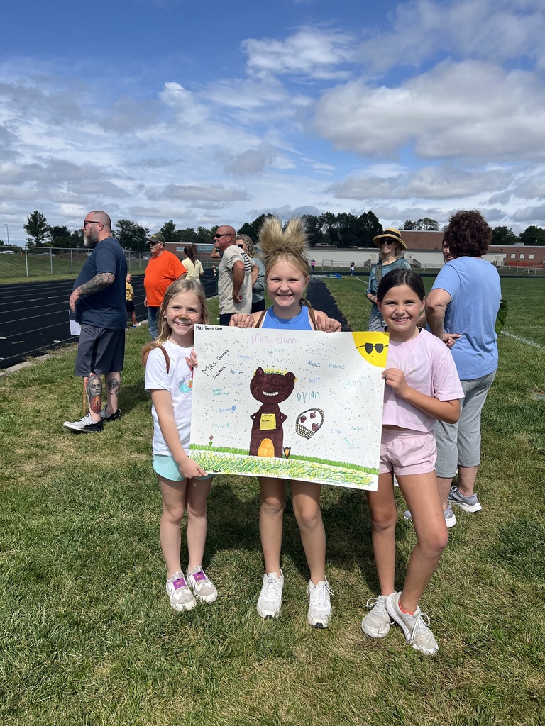 Three girls smiling holding up a poster of a squirrel kicking a soccer ball.