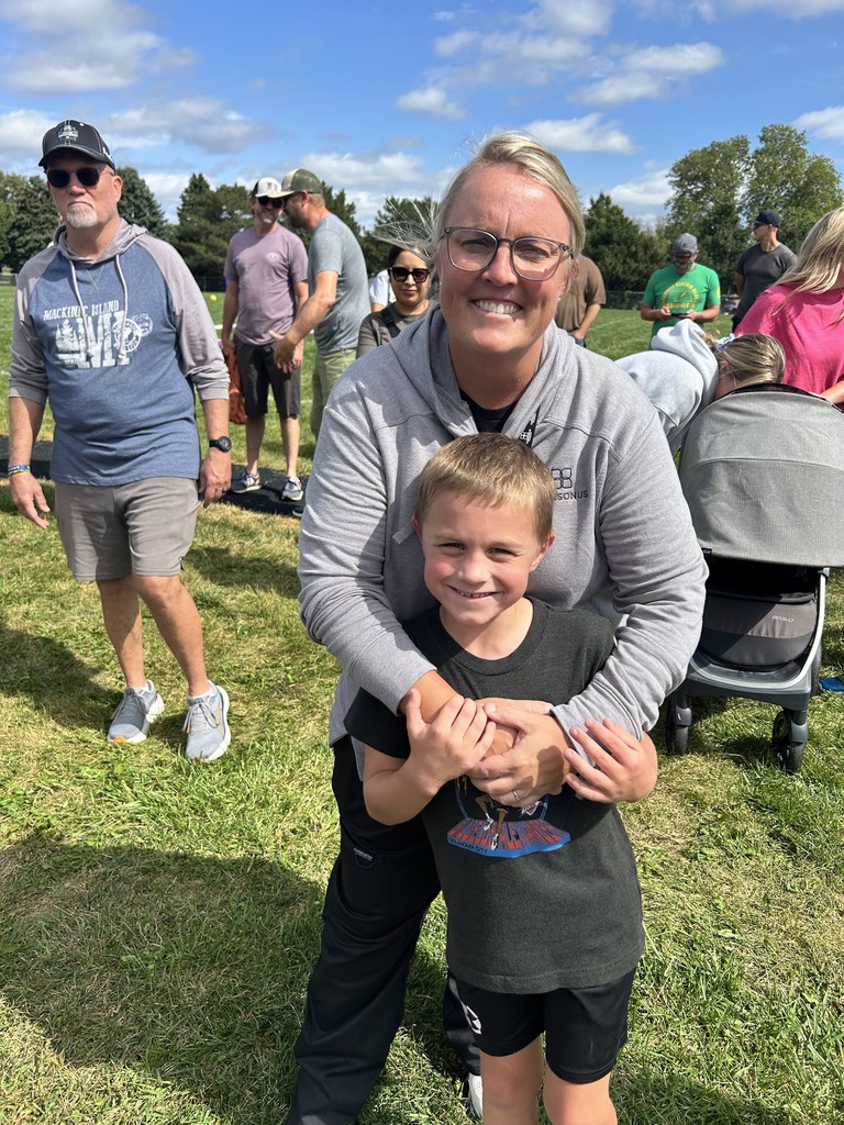 A boy being hugged by his mom on a grass field with other adults in the background.
