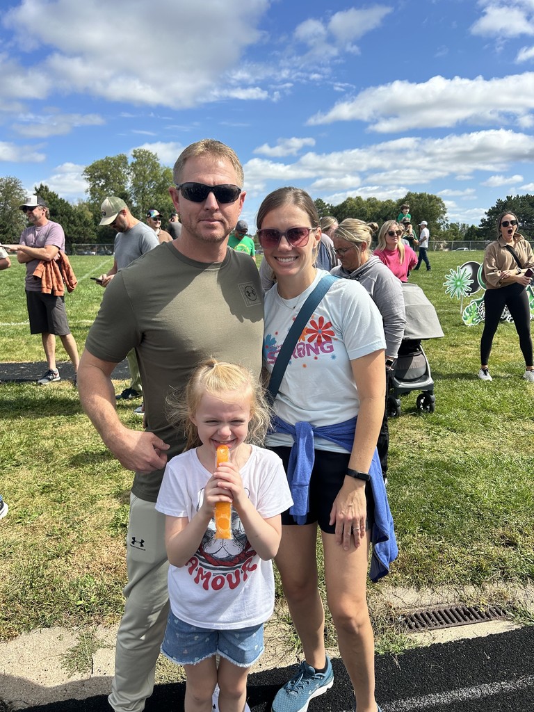 Girl standing holding an orange popsicle with her parents in front of a grass field.