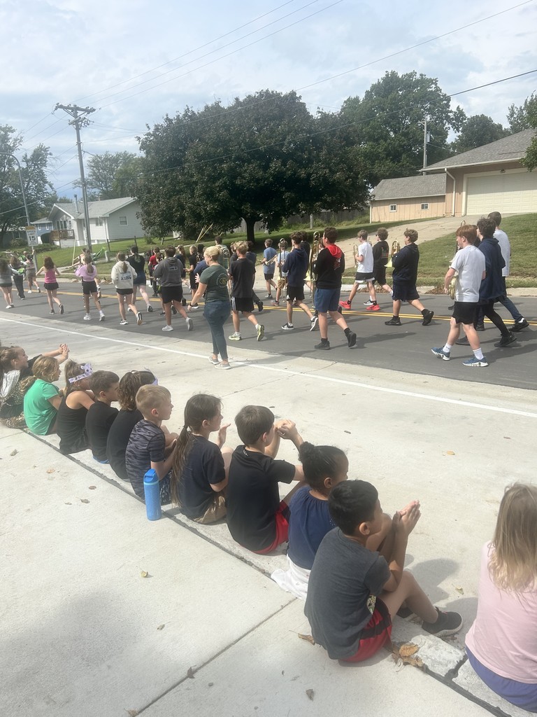 Marching band marching on a street with a group of students sitting on the sidewalk watching them.