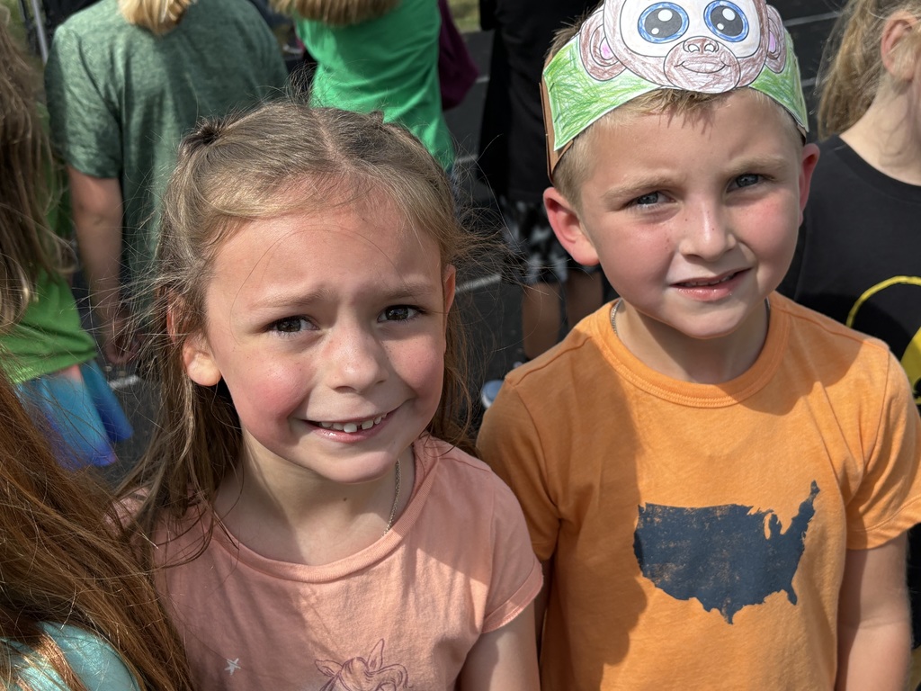 Two students standing wearing colorful shirts on a running track with one wearing a paper headband..