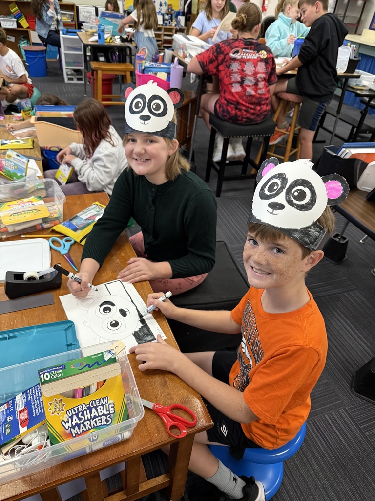 Two students sitting a table in a classroom with art supplies and a drawing of a panda visible.