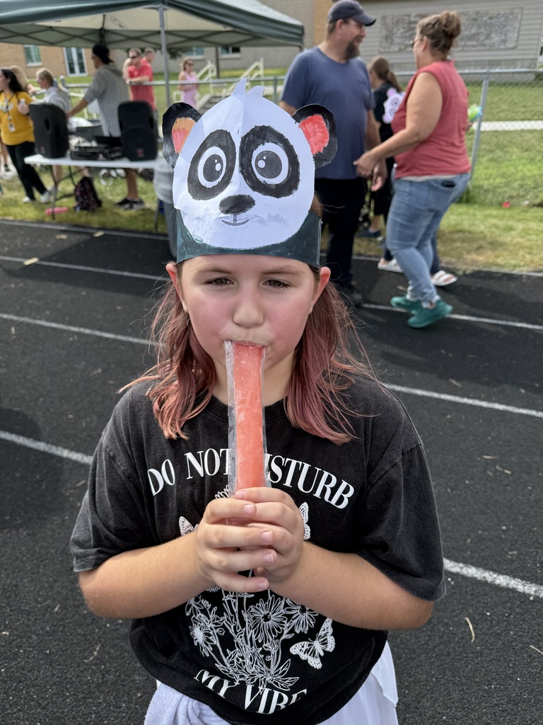 Girl standing on a running track while wearing a paper headband of a panda and eating a popsicle.