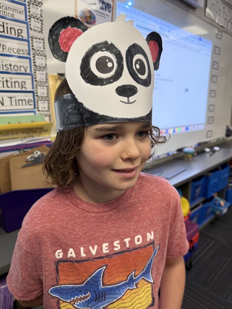 Boy standing in a classroom with a red shirt while wearing a panda paper head band.