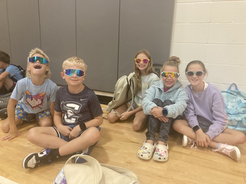 A group of 5 fourth graders sitting on the gym floor wearing sunglasses and smiling.
